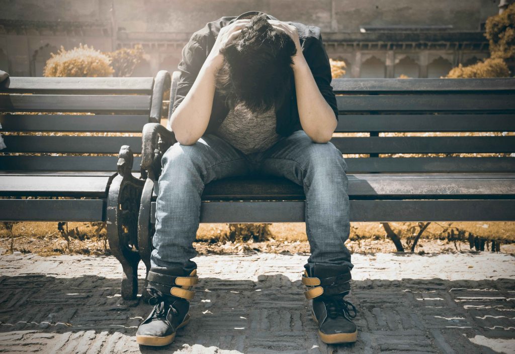 A man sitting on a park bench with his hands on his head and elbows on his knees. He looks depressed