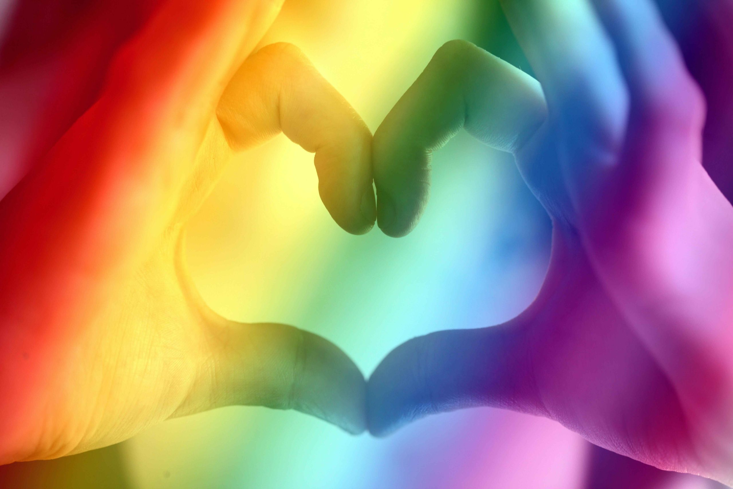 Two hands are forming the shape of a heart, and there is a rainbow glow over them.