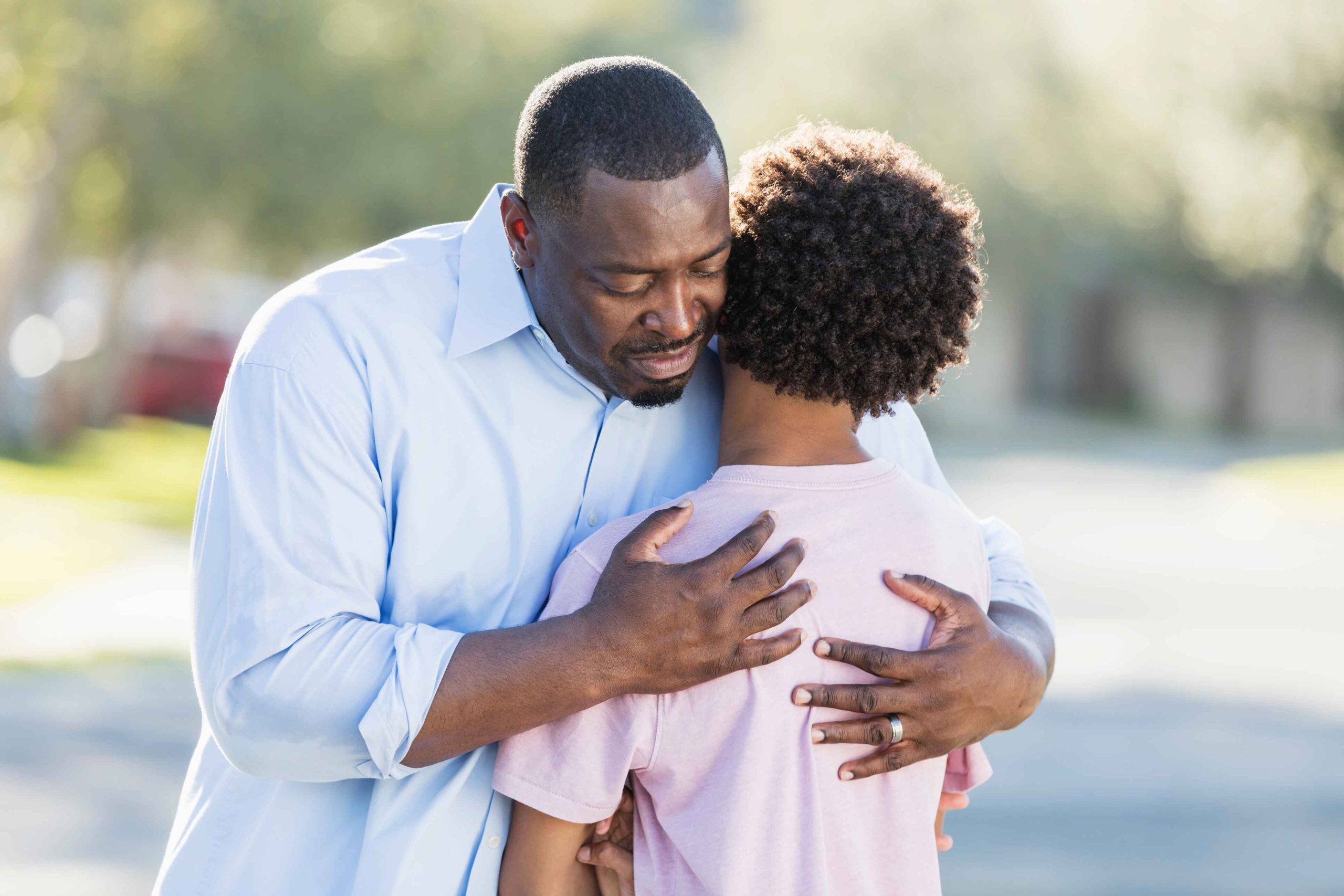 A man is hugging his son and his son's arms are crossed. The son seems like he is frustrated or sad.