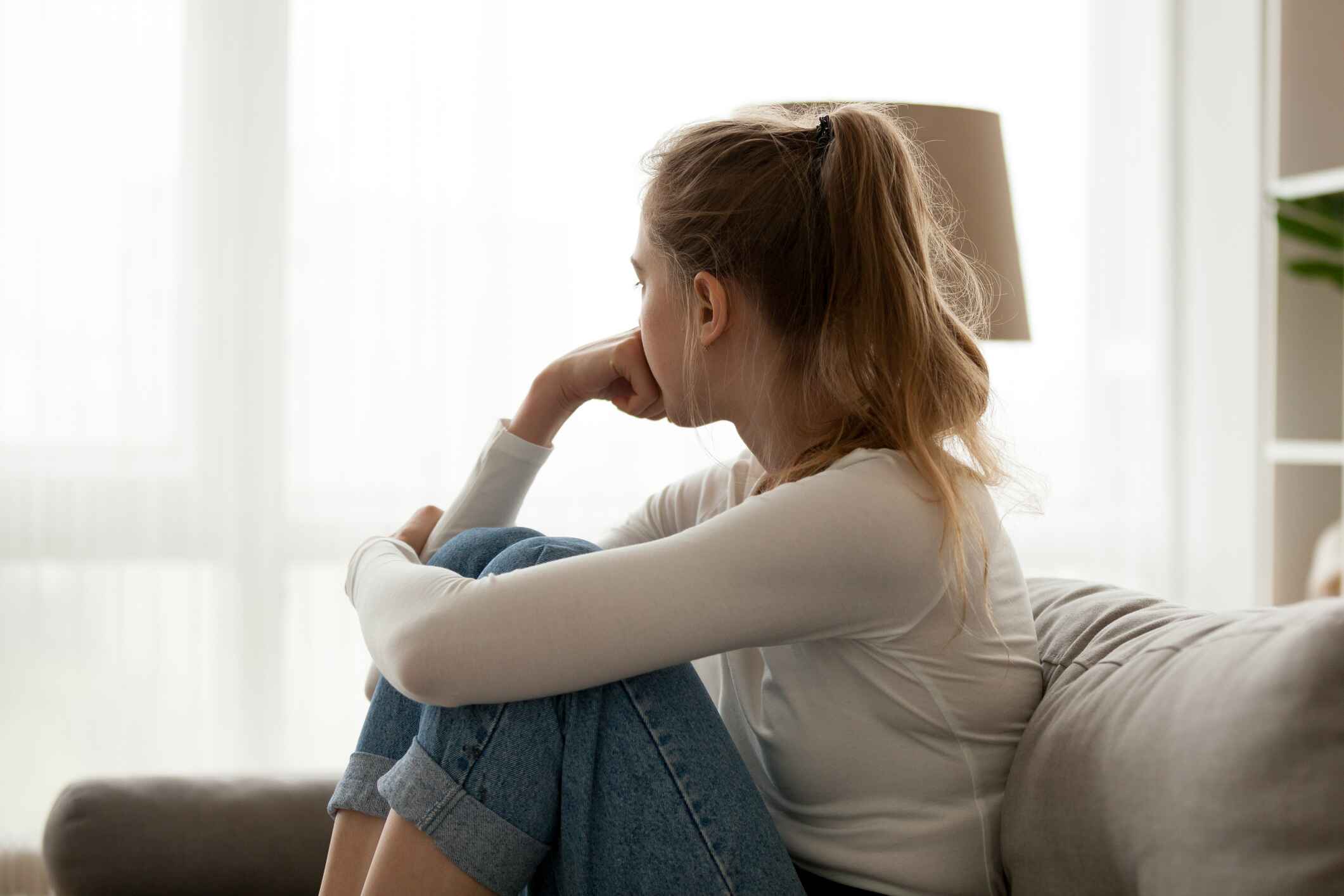 A girl sits on a couch with her arm wrapped around her knees. She's looking outside like she's sad.