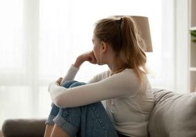 A girl sits on a couch with her arm wrapped around her knees. She's looking outside like she's sad.