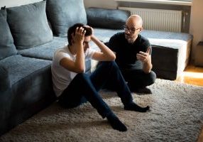 A depressed boy is sitting on the floor with his hands on his head. Another man is talking to him.