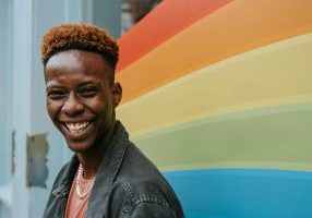 Smiling teenager with curly hair in front of rainbow wall.
