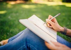 Person writing in journal, sitting in grass with jeans.
